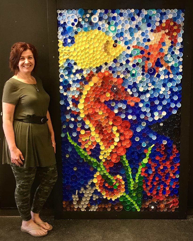 Art Teacher standing in from of a mosaic art piece. plastic caps in shapes of a fish, seahorse and lobster